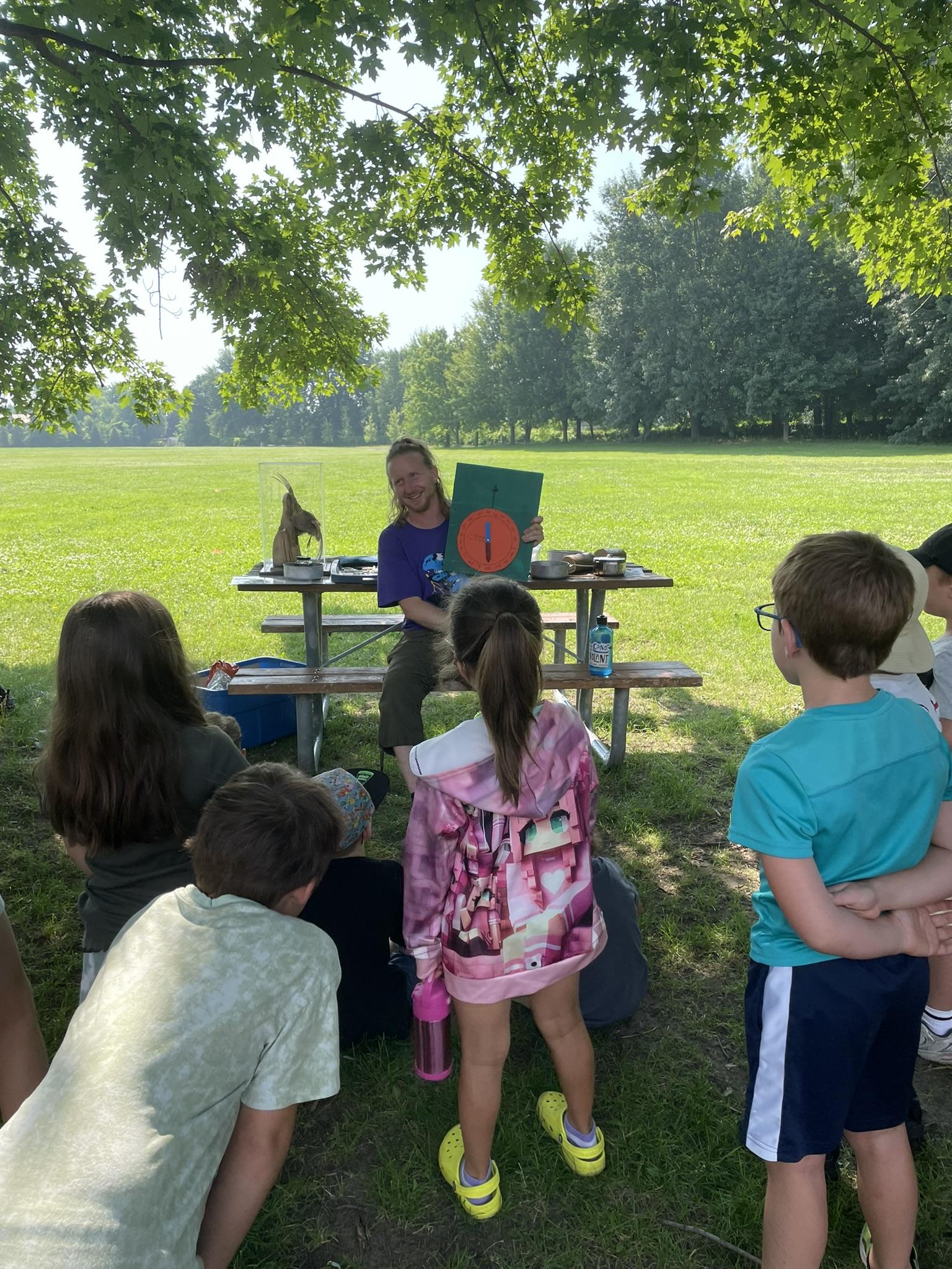 Une femme est assise à une table de pique-nique en plein air, tenant un livre et enseignant à un groupe d'enfants rassemblés autour d'elle à l'ombre des arbres par une journée ensoleillée.