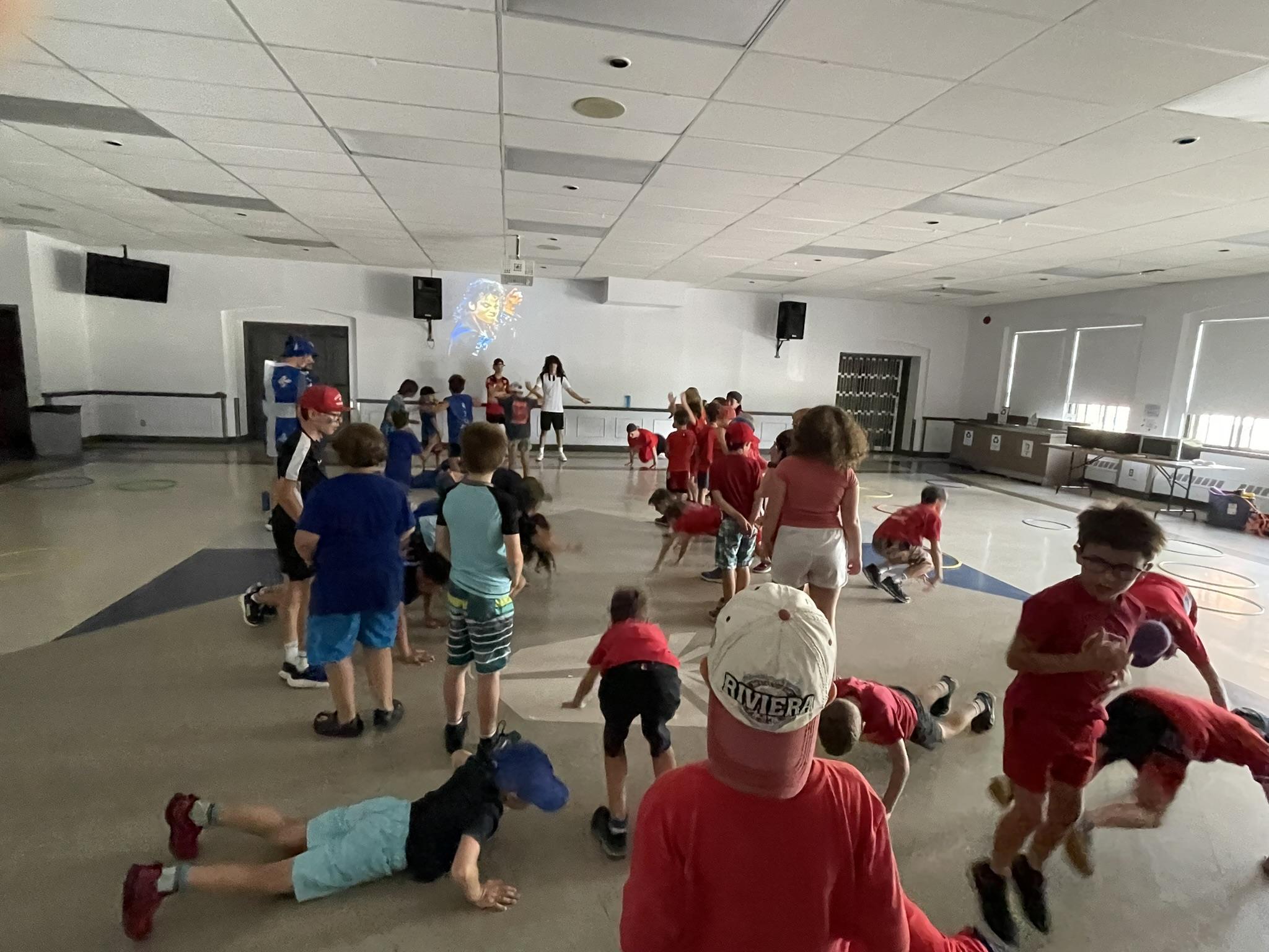 Dans une salle bien éclairée, un groupe d'enfants participe à des activités physiques, certains faisant des pompes et d'autres restant debout ou marchant. Un adulte supervise l'activité depuis l'arrière-plan, près d'un mur décoré d'une carte colorée.