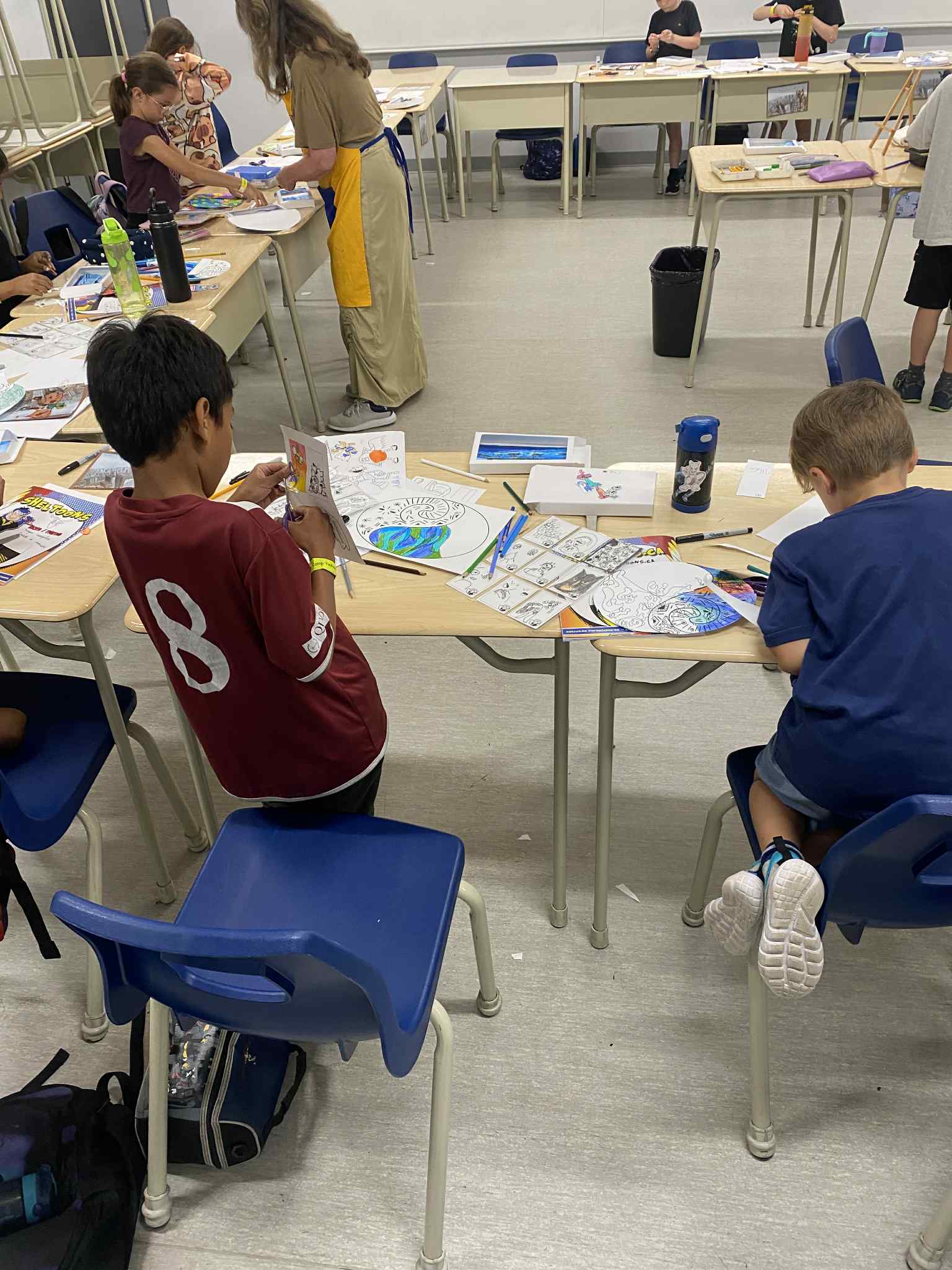 Des enfants assis à des bureaux dans une salle de classe, s'adonnant à des activités de dessin et de coloriage avec diverses fournitures artistiques étalées sur les tables. Un enseignant se tient à l'arrière-plan et la pièce dégage une atmosphère décontractée et créative.