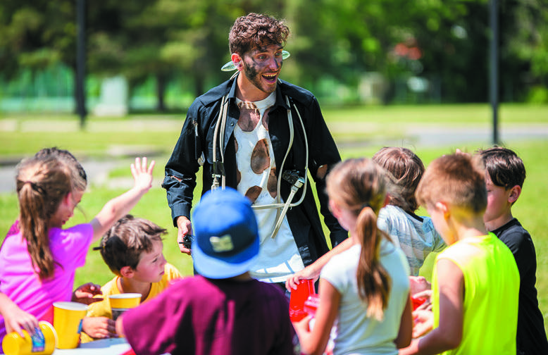 Un homme vêtu d'un drôle de costume avec de la suie sur le visage rit avec un groupe d'enfants qui jouent en plein air à une table avec des tasses colorées par une journée ensoleillée.