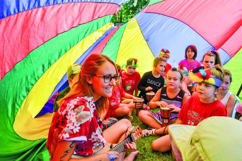 Une femme avec des lunettes et une guitare est assise sous un parachute coloré en plein air, entourée d'enfants portant des chemises rouges et des bandeaux de plumes, qui écoutent tous attentivement. La scène est vivante et lumineuse.